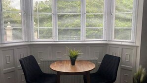 Ein Erker mit weißen Fensterläden, ein runder Holztisch und zwei schwarze Stühle.