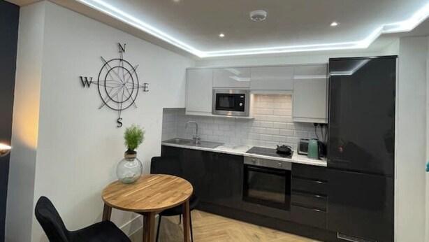 Eine moderne Küche mit schwarzen Schränken, einem Edelstahlkühlschrank, einer Mikrowelle und einem Spülbecken. An der Wand hängt eine Kompassdekoration und es steht ein kleiner Tisch mit Stühlen bereit.