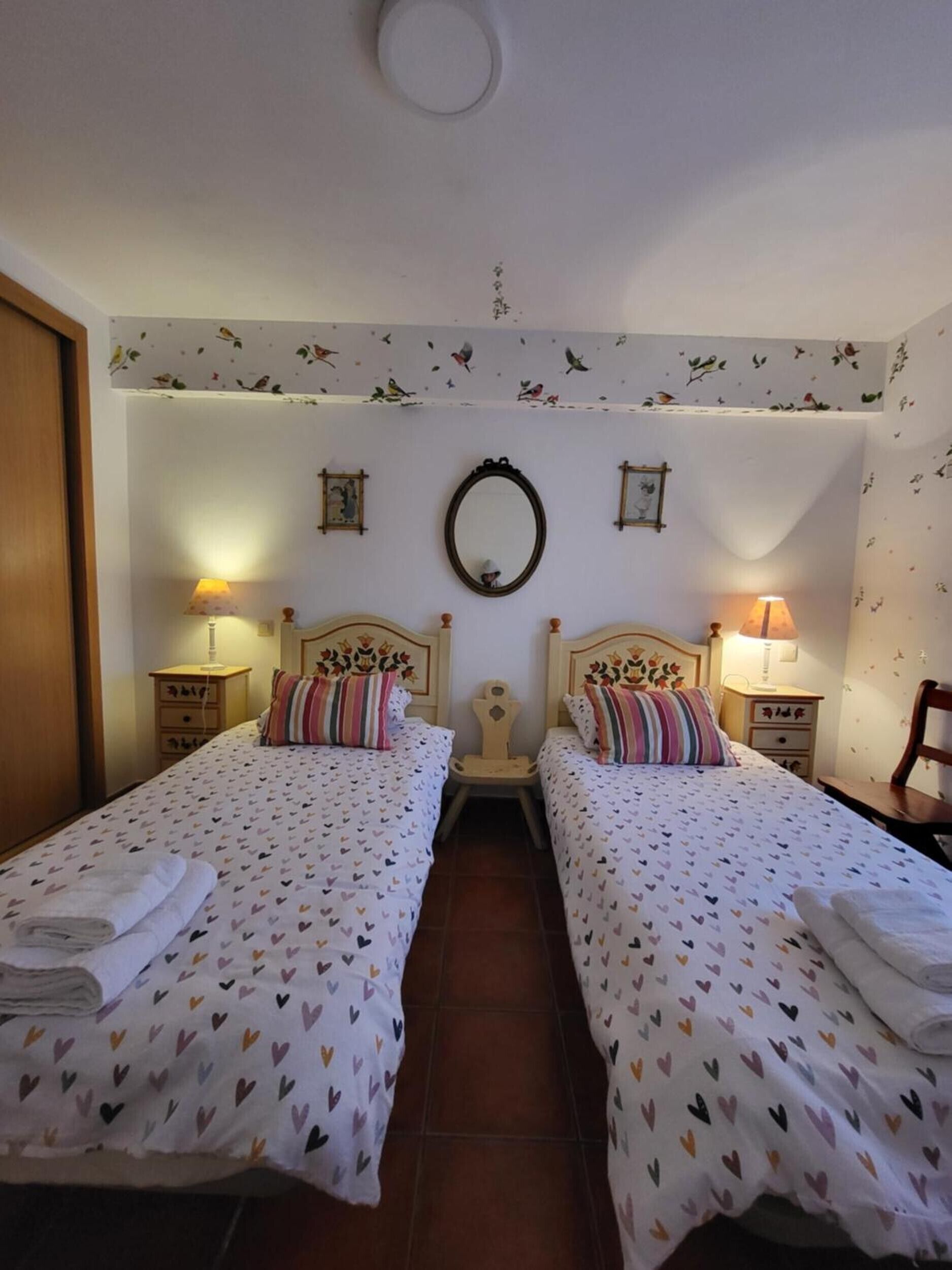 A room with two beds, each with a heart-patterned bedspread, a mirror on the wall, and two bedside lamps.