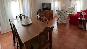 Un coin repas avec une table et des chaises en bois, un téléviseur posé sur un meuble, et un canapé rouge à l’arrière-plan.