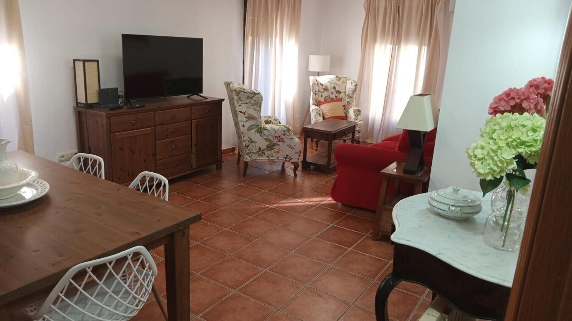 Un salon comprenant un téléviseur, un fauteuil à motifs floraux, un canapé rouge, une table à manger avec des chaises et un vase rempli de fleurs.