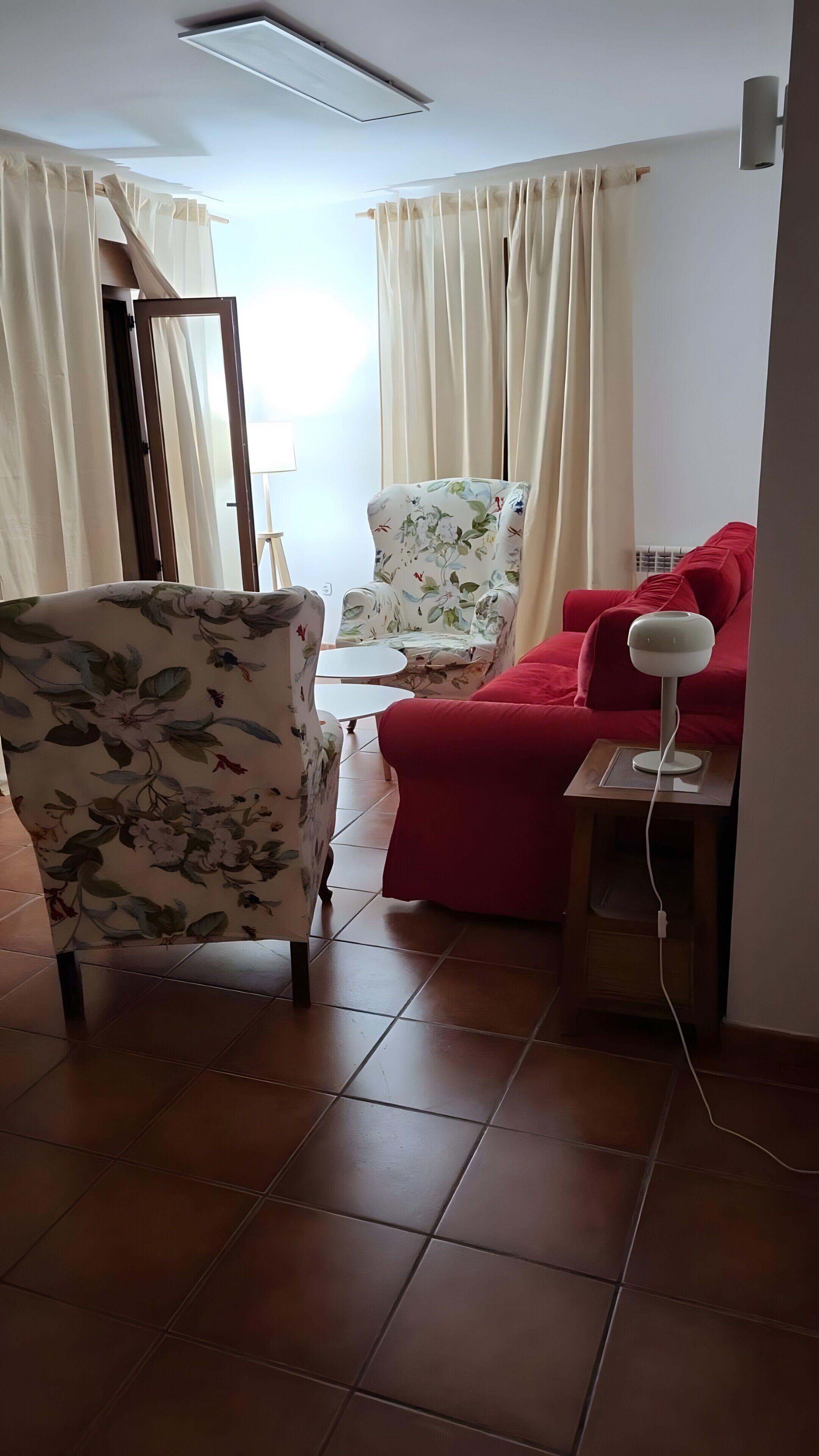 Een woonkamer met een fauteuil met bloemenprint, een rode bank, een bijzettafeltje en een staande lamp.