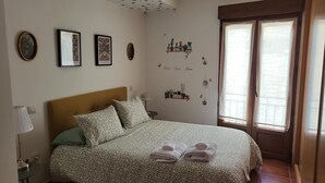Une chambre à coucher comprenant un lit, des oreillers, une table de chevet, une lampe, une fenêtre avec des rideaux et des éléments de décoration murale, notamment des tableaux encadrés et un motif décoratif adhésif.