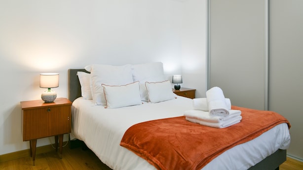 Un lit bien fait, avec une literie blanche et un plaid orange, une lampe de chevet et une table de chevet en bois.