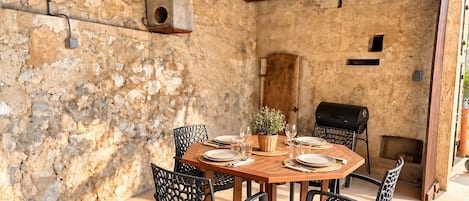 An outdoor dining area with a rustic stone wall, wooden ceiling beams, and a round wooden table with chairs.