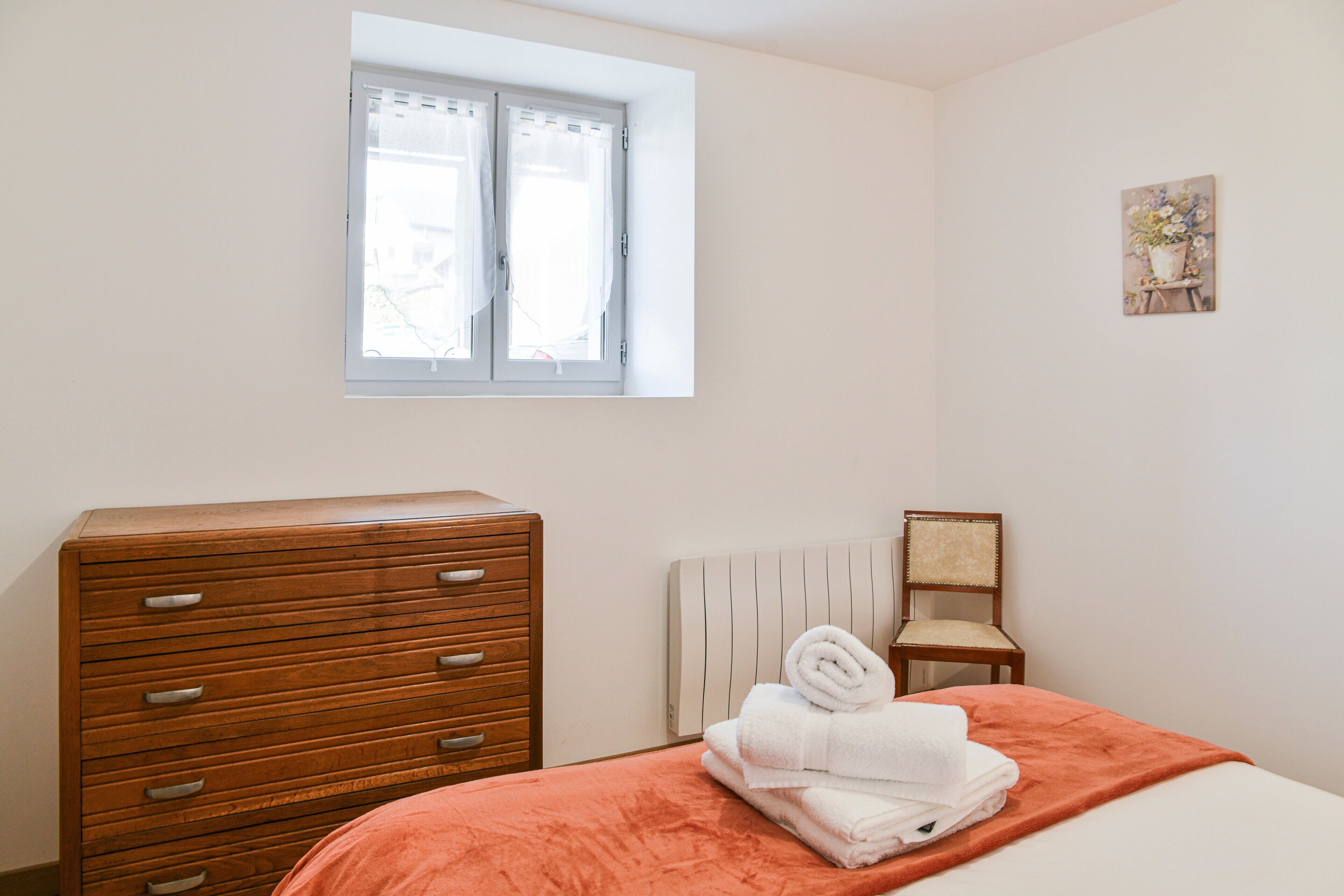 A bedroom with a wooden dresser, a bed with an orange blanket, a chair, and a window with curtains.