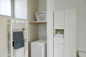 Un espace buanderie comprenant un sèche-linge blanc, un porte-serviettes blanc fixé au mur et un panier à linge blanc posé sur une étagère.