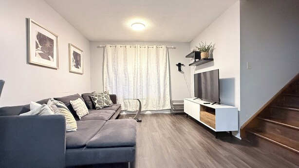 Een moderne woonkamer met een grijze bank, een flatscreen televisie op een witte dressoir, en een trap aan de rechterkant.