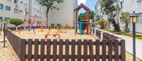 A playground with swings and climbing structures, surrounded by a wooden fence and pebbles.