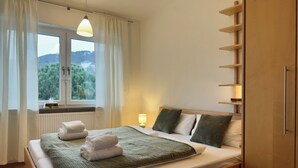A neatly made bed with a green blanket and white towels, a bedside lamp, and a view of trees through a window with white curtains. - Detached single-family home above the rooftops of Bad Dürkheim (Bad Dürkheim)