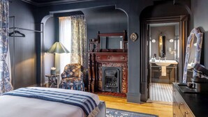 A bedroom with a bed, a chair, a fireplace, a mirror, and a bathroom visible through an archway.