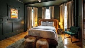 A bedroom with a large bed, two bedside lamps, a green chair, and a stained glass window. - The Robert Frost - Ash Street Inn (Manchester)