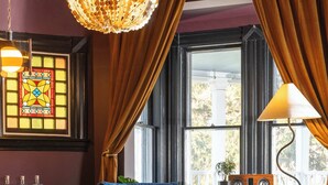 A room with a blue armchair, a round table, a chandelier, a stained glass window, and a view of the outdoors. - The Robert Frost - Ash Street Inn (Manchester)