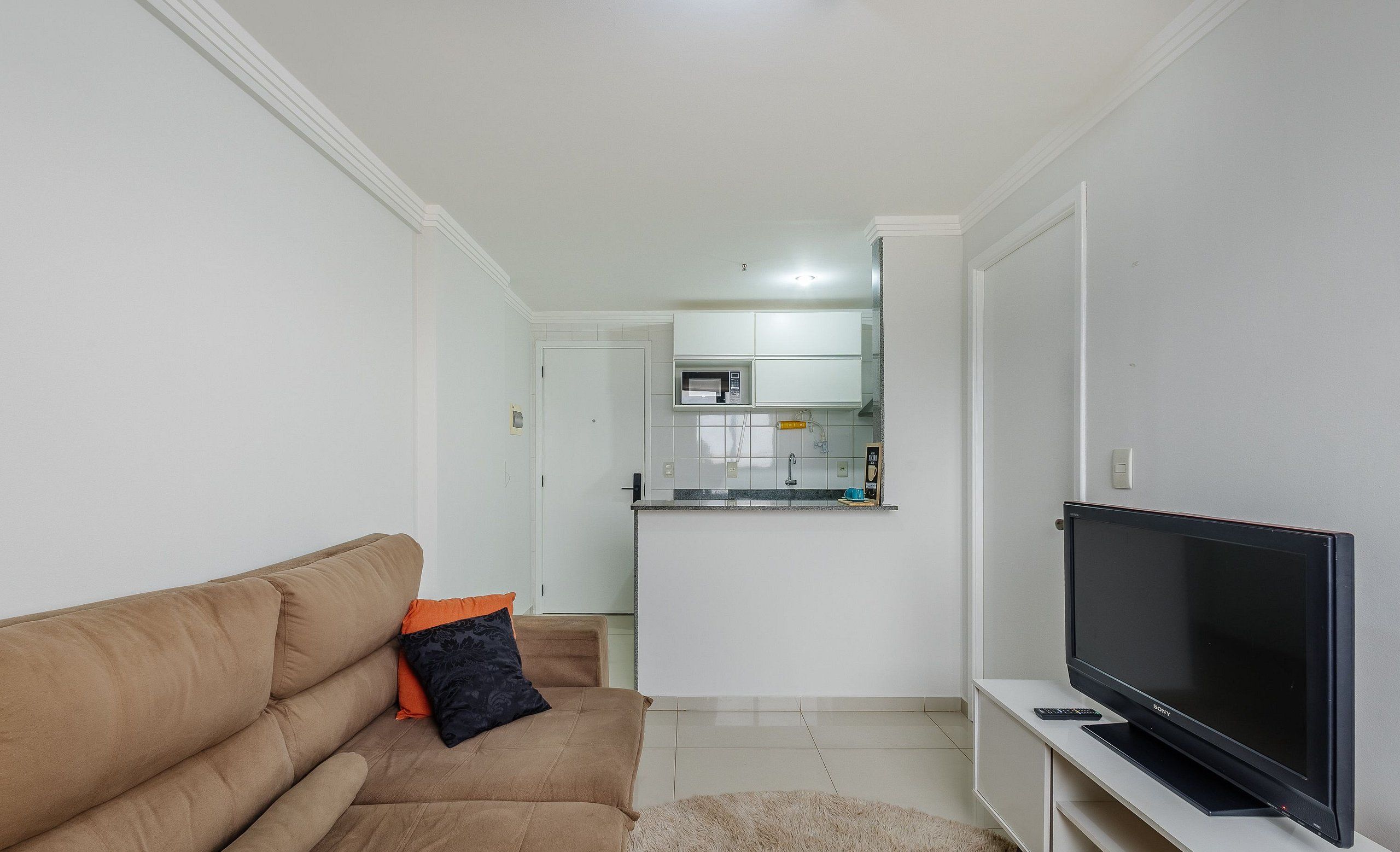A living room with a beige sofa, a flat-screen TV, and a kitchen in the background.