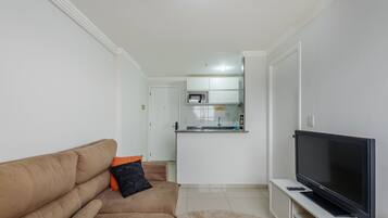A living room with a beige sofa, a flat-screen TV, and a kitchen in the background.