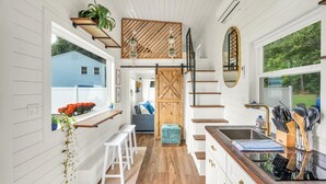 Eine kleine Küche mit weißen Schränken, einer Holztreppe und Blick auf ein blaues Haus.