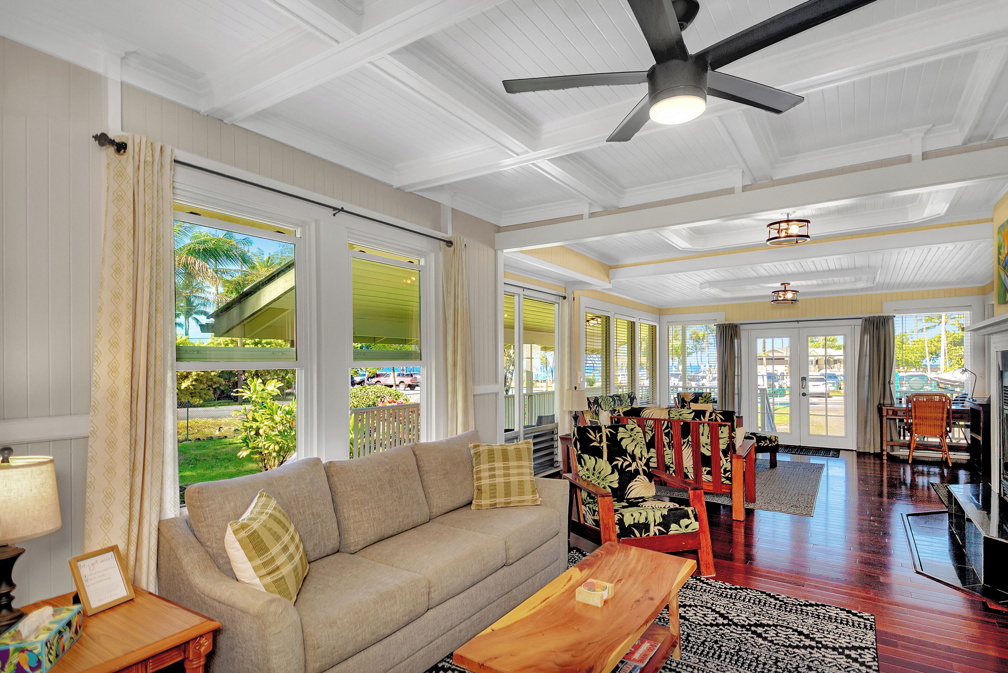 A spacious living room with a sofa, coffee table, and chairs, large windows with curtains, and a ceiling fan.