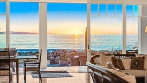 A room with a large window overlooking the sea at sunset, furnished with wicker chairs and a sofa.