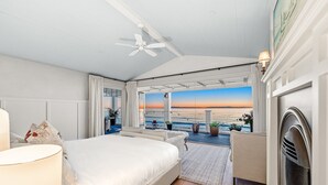 A bedroom with a large bed, a ceiling fan, a fireplace, and a view of the ocean.