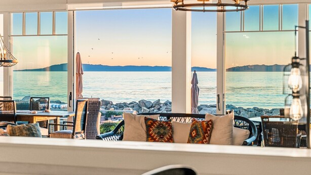 A seaside view from a room with large windows, wicker furniture, and a hanging light fixture.