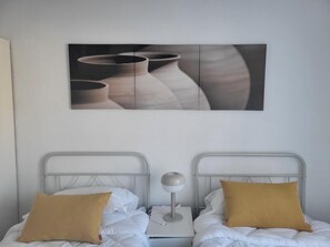 Ein Zimmer mit zwei Betten, jeweils mit Metallgestell und weißer Bettwäsche, einem Nachttisch mit Lampe sowie einem großen, abstrakten Gemälde an der Wand.