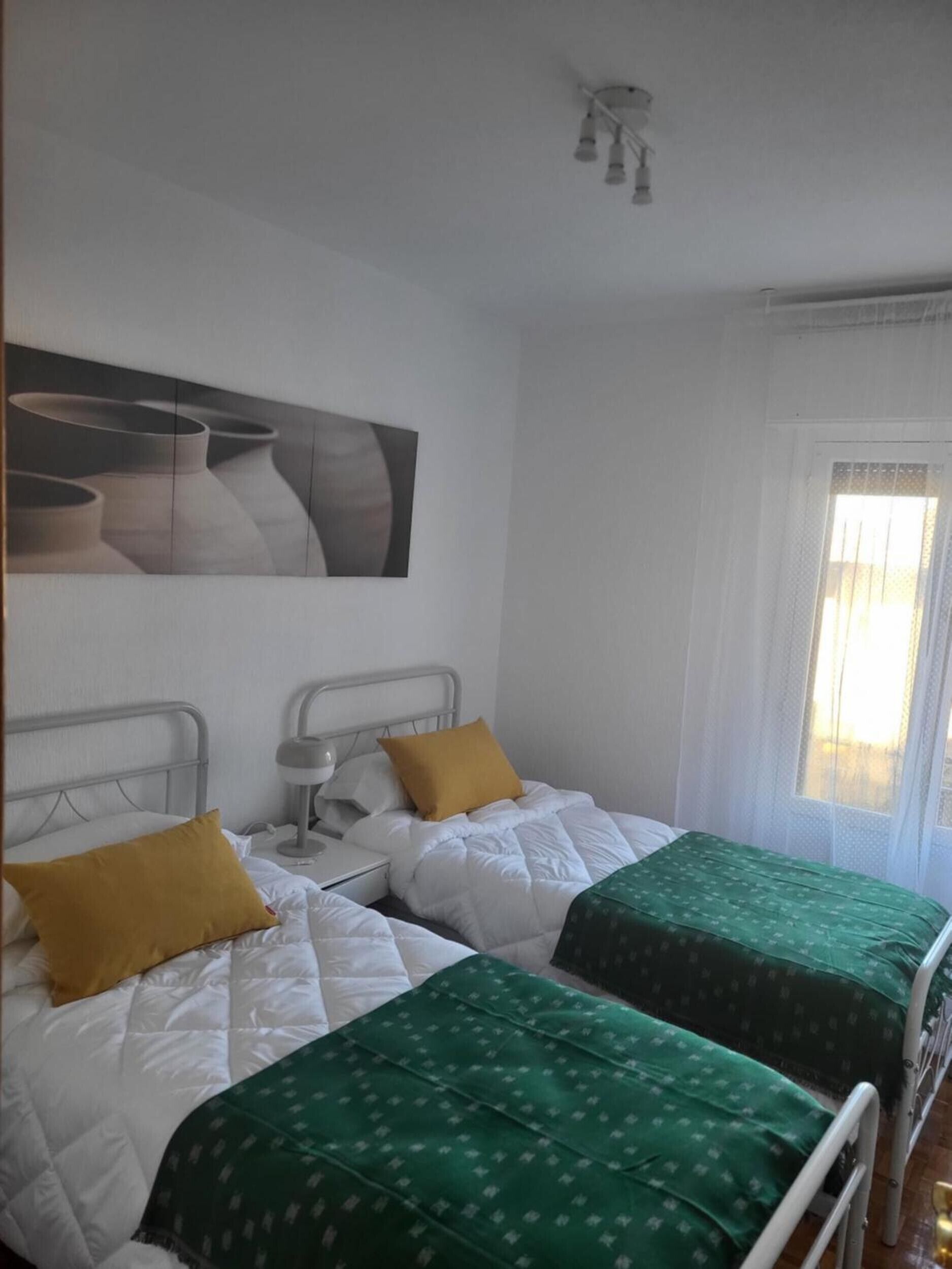 A room with two beds, each with a distinct green and yellow bedspread, a bedside table with a lamp, and a wall-mounted artwork.