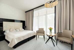 Ein Hotelzimmer mit einem Bett, zwei Sesseln, einem kleinen Tisch, einer Vase mit Blumen und einem großen Fenster mit Vorhängen.