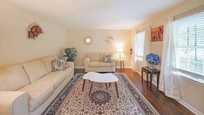 Ein Wohnzimmer mit einem beige farbenen Sofa, einem gemusterten Teppich, einem kleinen weißen Couchtisch, einem Beistelltisch mit einer Lampe und einem Fenster mit Jalousien.