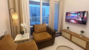 Un espacio habitable compacto con un sofá, una pequeña mesa con una planta, un televisor montado en la pared y un balcón con vistas a edificios altos.