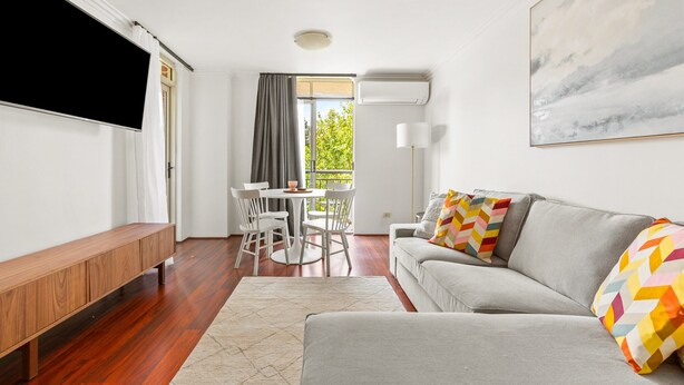 A modern living room with a grey sofa, colorful pillows, a wooden bench, a dining table with chairs, and a large painting on the wall. - Green Views & Great Location: Inner-city 1-bed (Sydney)