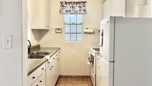 Eine kleine Küche mit weißen Schränken, einem eingebauten Ofen, einem Kühlschrank, einem Fenster mit gemusterten Vorhängen und einem Deckenventilator.