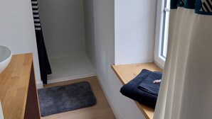 A small, narrow room with a wooden floor, a white wall, a window with patterned curtains, a wooden bench, and a black towel on the bench. - Ty PenAr Pont étage (Saint-Pol-de-Léon)