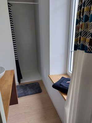 Une petite pièce étroite avec un plancher en bois, un mur blanc, une fenêtre ornée de rideaux à motifs, un banc en bois et une serviette noire posée sur le banc.