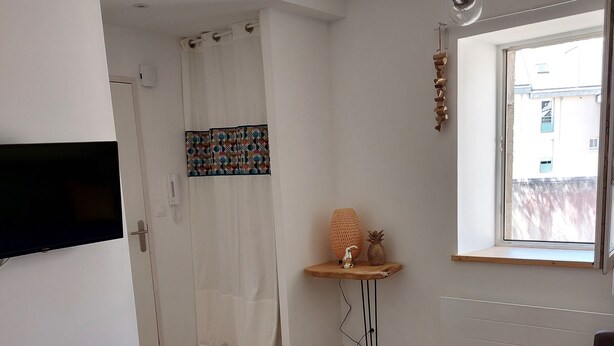 A modern, well-lit room with a dining area, a TV mounted on the wall, a window with a view of buildings, and a door with a curtain. - Ty PenAr Pont étage (Saint-Pol-de-Léon)