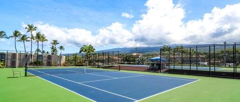Una cancha de tenis con una red, rodeada por una valla, palmeras y montañas al fondo.