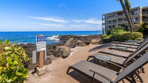 A beachside view with lounge chairs, a sign indicating beach access times, and a building with balconies overlooking the ocean. - Great Location AC & Wifi Concierge Included! (Kailua-Kona)