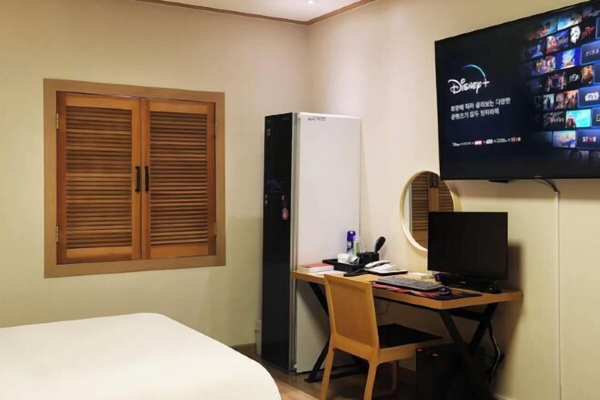A hotel room with a bed, a desk with a computer, a TV mounted on the wall, and a refrigerator.