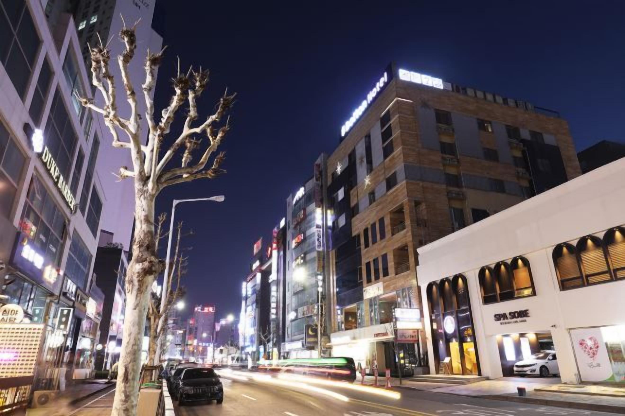 Eine beleuchtete Stadtstraße bei Nacht mit Gebäuden, einem Baum und einem fahrenden Auto.