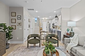 Una sala moderna con un sofá, dos sillones, una mesita de centro y una zona de comedor con una mesa y sillas. Se observa una cocina al fondo con armarios y un microondas.
