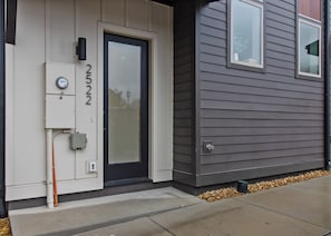Una casa moderna con revestimiento gris en el exterior, una puerta principal negra y un cuadro eléctrico visible en el lado izquierdo.