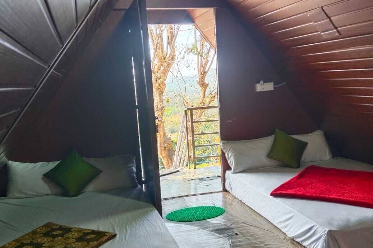 A room with two beds, a wooden ceiling, and a view of trees outside.