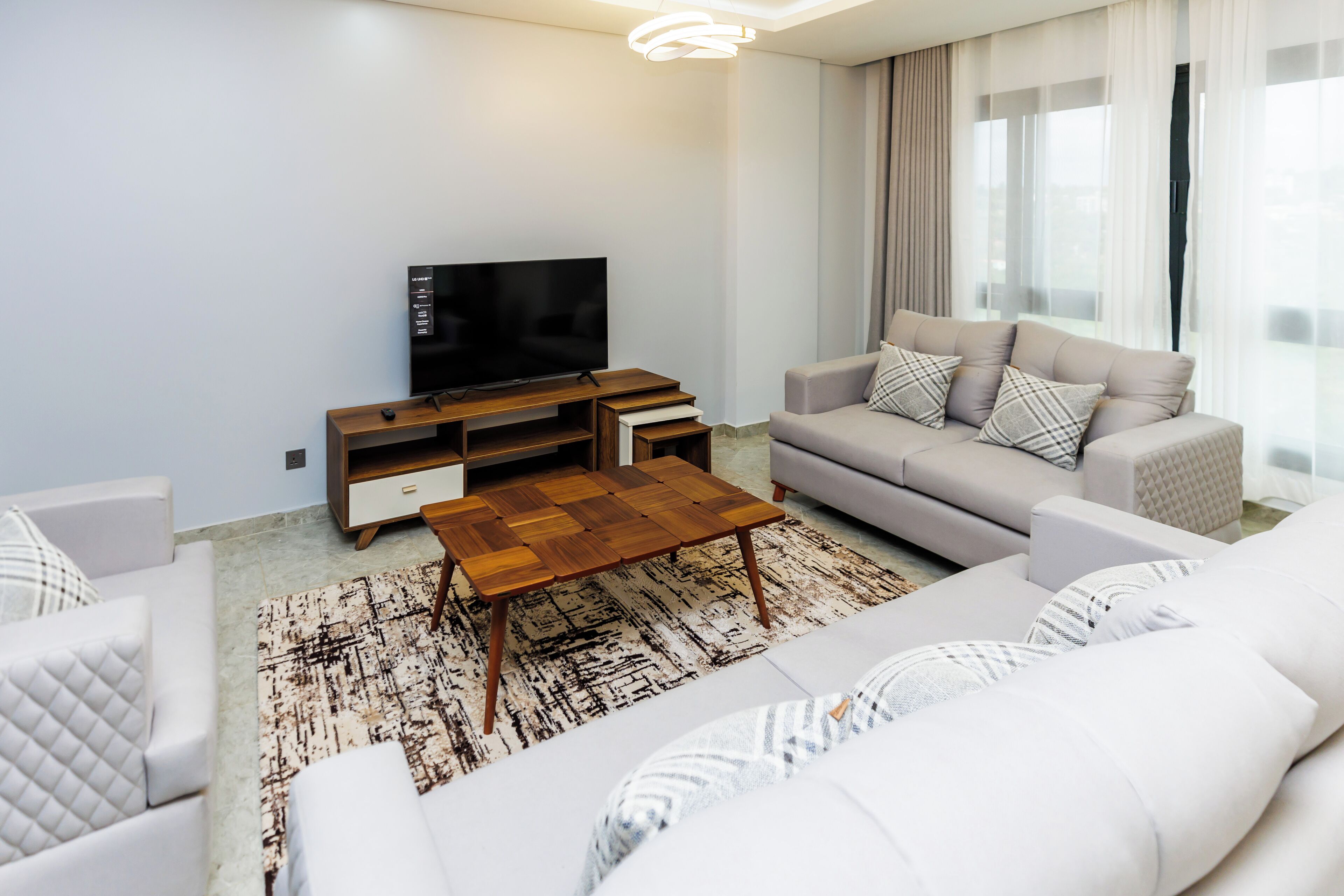 A modern living room with a flat-screen TV, a wooden coffee table, a grey sofa, and a patterned rug.