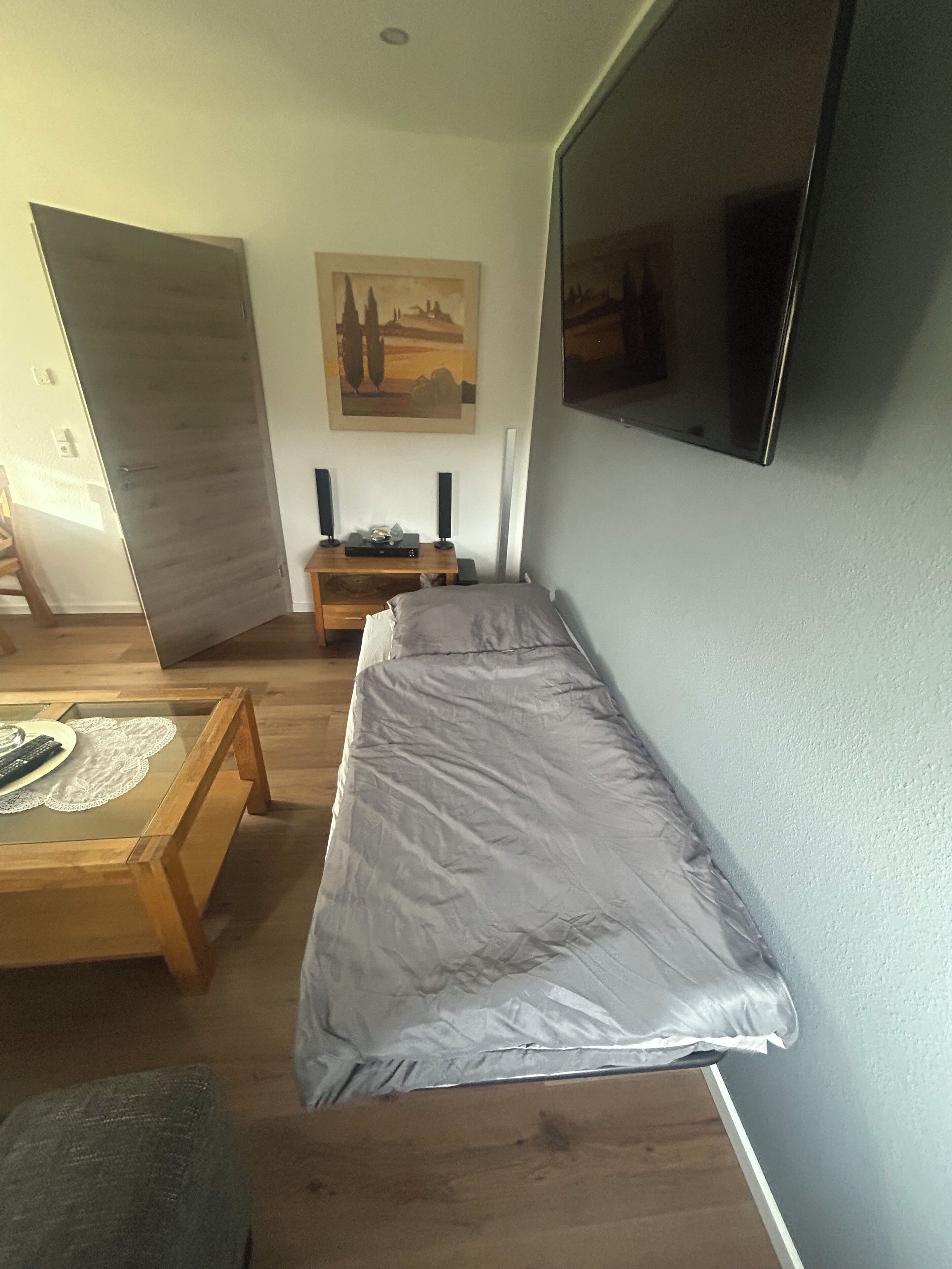 Un espacio habitable pequeño con una cama, una mesita de centro de madera, una mesita auxiliar y un televisor de pared.