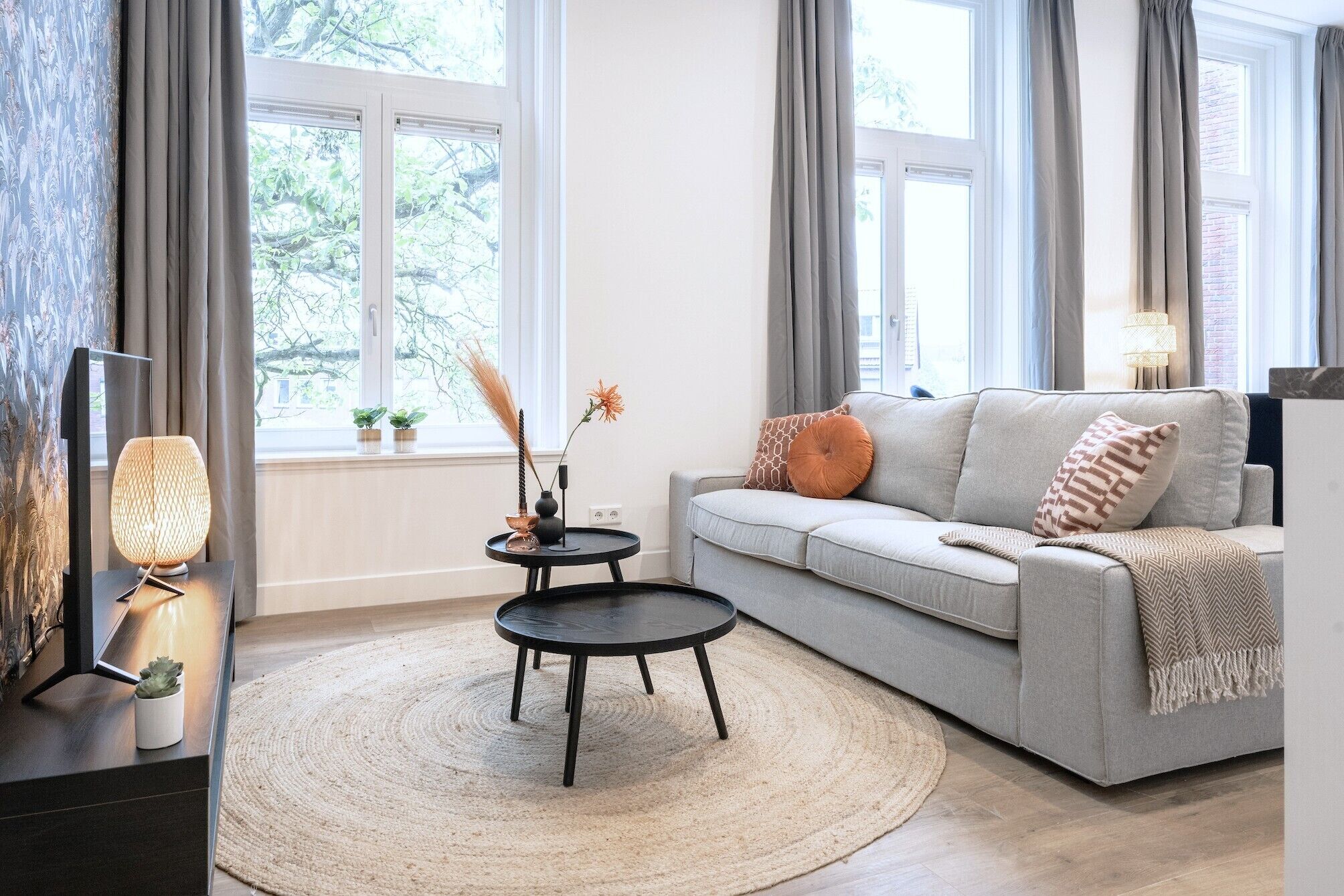 A modern living room with a grey sofa, a round coffee table, and a TV on a stand. There are two windows with curtains, a lamp on a side table, and a small plant on the windowsill.