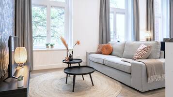A modern living room with a grey sofa, a round coffee table, and a TV on a stand. There are two windows with curtains, a lamp on a side table, and a small plant on the windowsill.