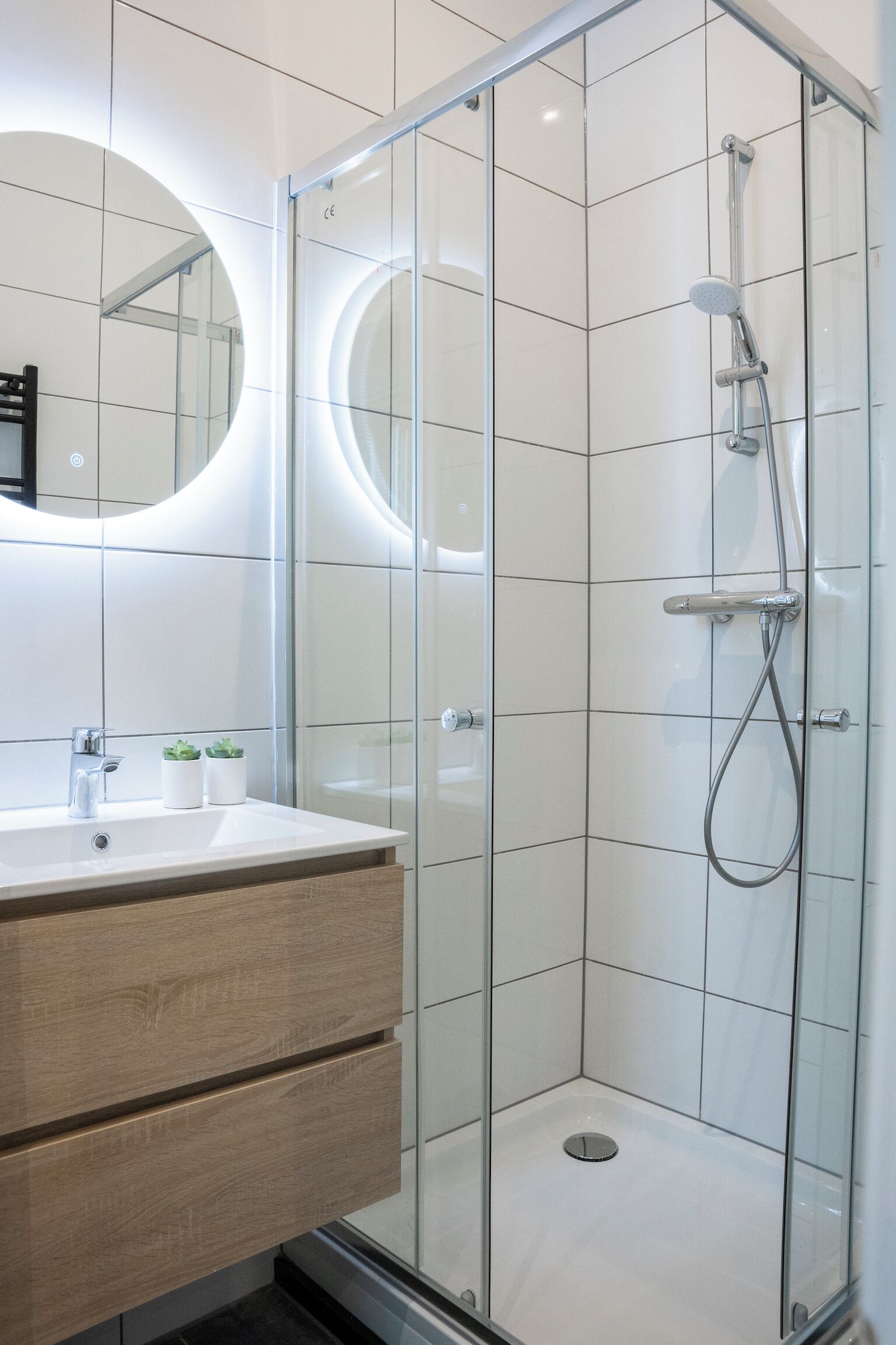 Un baño moderno con cabina de ducha de vidrio, un espejo redondo y un mueble de tocador de madera con cajones.