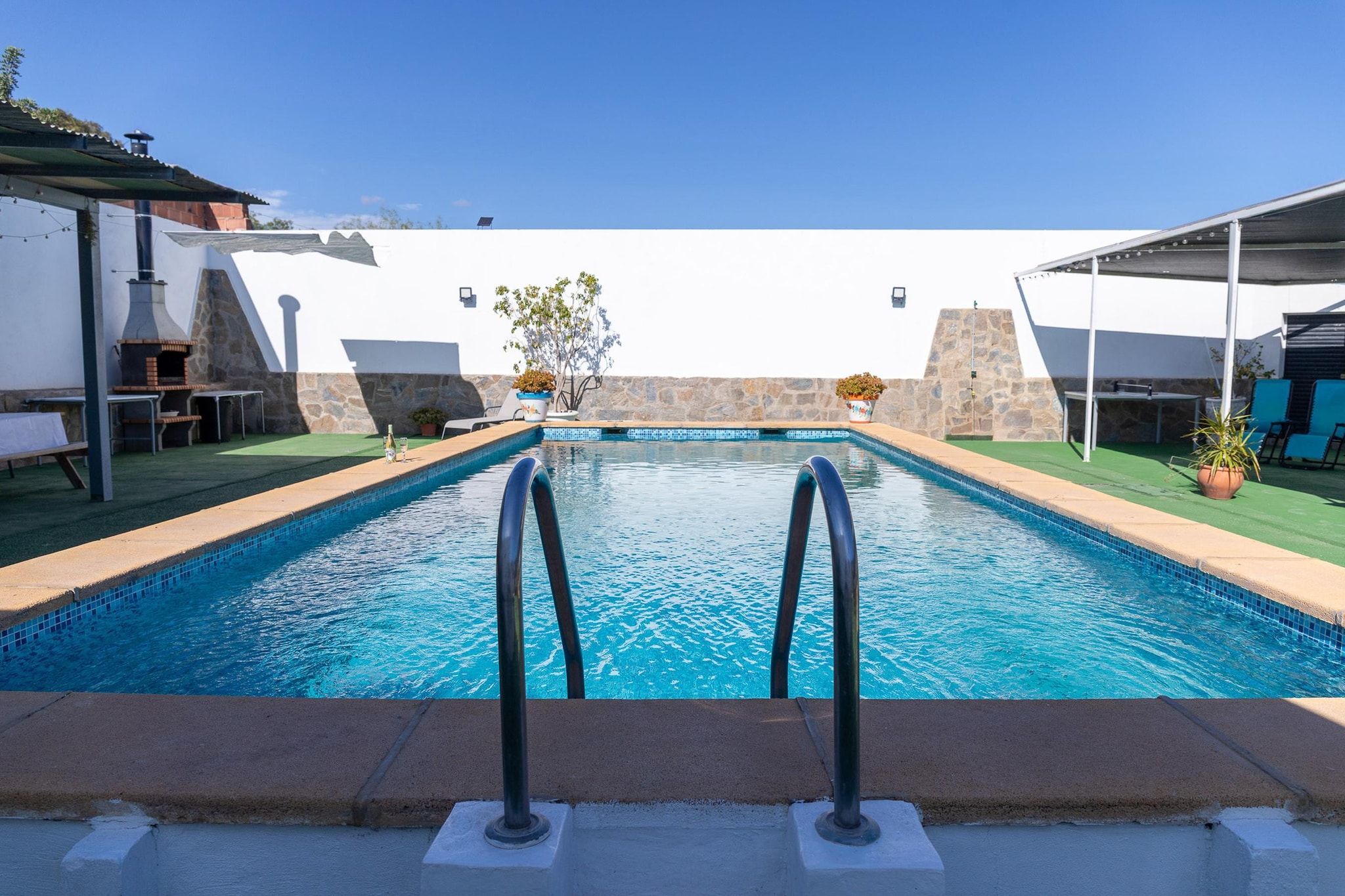 Une piscine bordée d’un mur de pierre, avec un ciel bleu et dégagé.