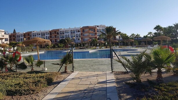 A swimming pool surrounded by palm trees and a fence, with buildings in the background. - Perfect Getaway in Vera (Almeria)