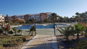 A swimming pool surrounded by palm trees and a fence, with buildings in the background. - Apartment near Vera Naturist Beach (Almeria)