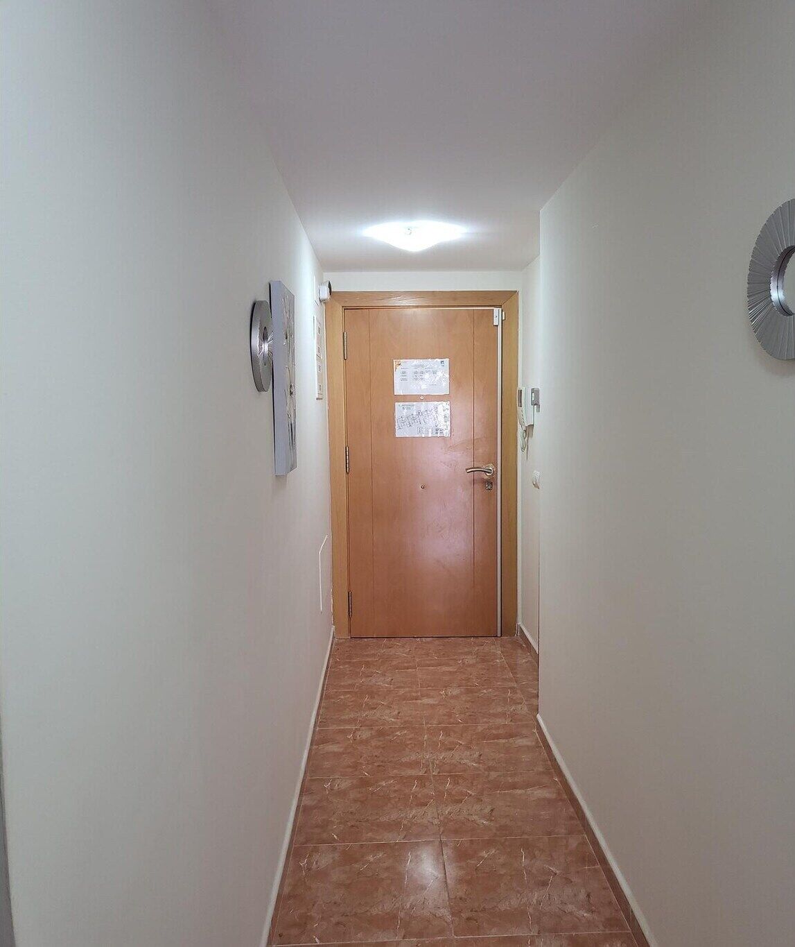 Un couloir étroit, avec une porte en bois au fond, un luminaire au plafond et une horloge murale sur le côté droit.